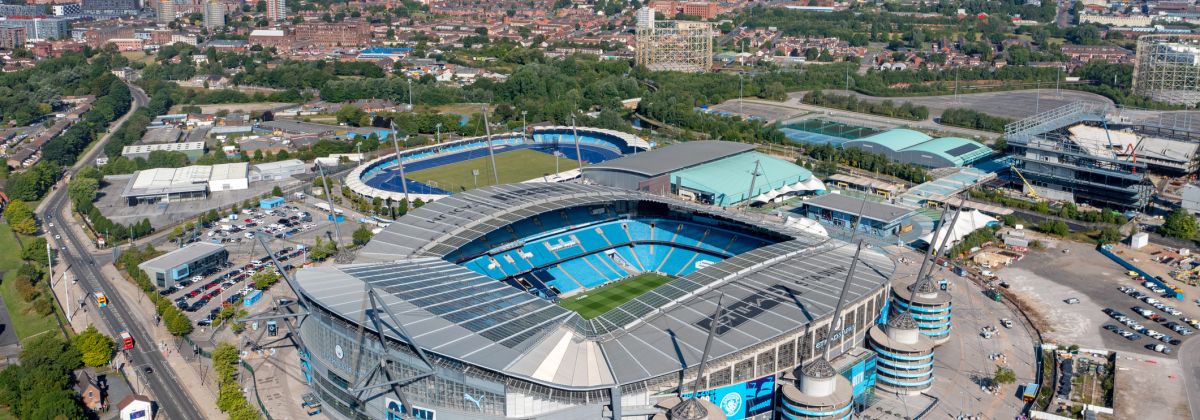 Aerial view of Etihad Stadium in Manchester, surrounded by parking lots, training grounds, and city buildings in the distance.